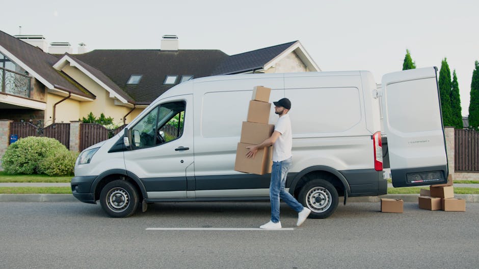 A man wearing a white t-shirt, blue jeans, white sneakers, and a black cap is loading three cardboard boxes into the side of a white cargo van parked on a residential street. The van, which belongs to Finchley Man and Van, features black accents and an open rear door. Several additional boxes are placed on the pavement nearby. The background shows modern houses with sloped roofs, chimneys, and well-maintained gardens, with some greenery and a wooden fence visible. The image captures the loading process during daytime, illustrating home relocation and furniture transport activities as part of a professional removals service.