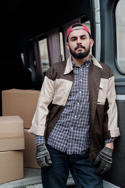 A man and a woman are standing beside an open delivery van parked on a paved area, involved in a home relocation process. The man, dressed in a white polo shirt, black cap, and wearing wireless earbuds, is holding a clipboard and a pen, inspecting or recording details related to the boxes. The woman, wearing a white cap, black sleeveless top, and jeans, is holding a small cardboard box, preparing for the next step in packing or unloading. Inside the van, numerous stacked cardboard boxes of various sizes are visible, some sealed with packing tape, and others awaiting transport. The interior of the vehicle is dark, contrasting with the outdoor natural lighting illuminating the scene. Moving equipment such as a trolley or straps is not visible. The setting showcases a professional house and flat removal service by Finchley Man and Van, emphasizing furniture transport, packing, and loading processes essential to residential relocation services.