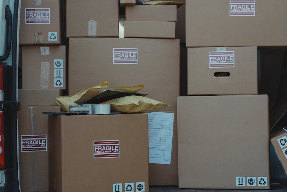 A collection of cardboard boxes of various sizes, some labeled with 'FRAGILE HANDLE WITH CARE' stickers, are loaded into the back of a van, ready for a home relocation. The boxes are stacked neatly on