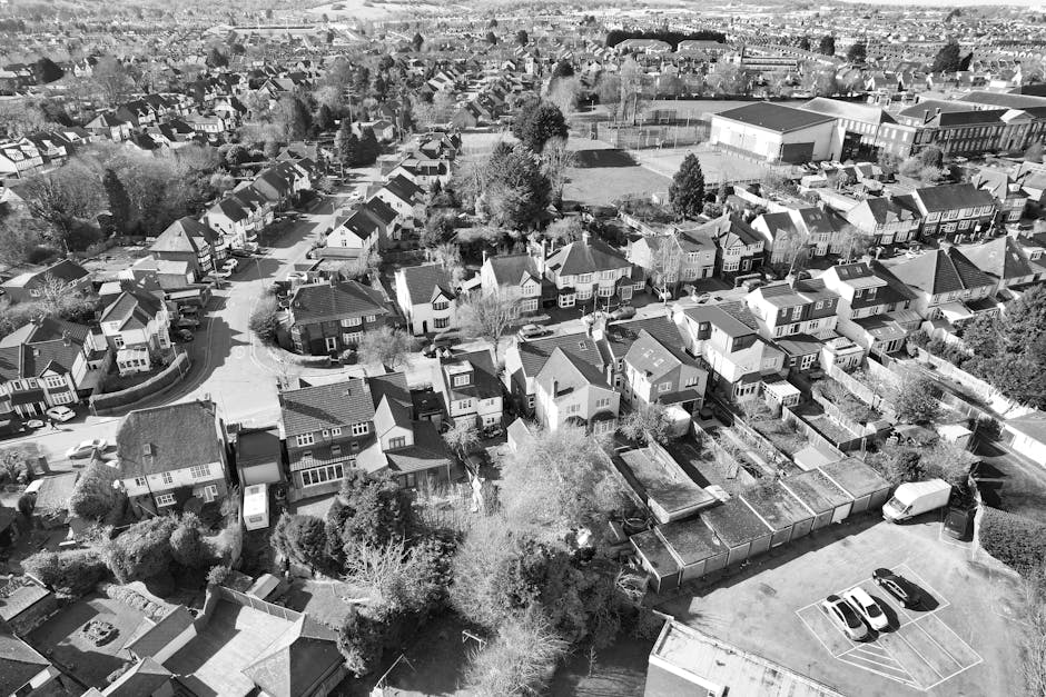 An aerial black and white photograph of a residential neighbourhood showing a variety of detached and semi-detached houses with pitched roofs, arranged along curved and straight streets. Several houses have visible front gardens, driveways, and small backyards enclosed by fences or walls. A few trees and shrubs are scattered throughout the area. In the foreground, a paved parking area contains three parked cars. Adjacent to this, there is a row of garages or storage sheds with flat roofs, possibly used for household storage during a house removal or moving process. The street scene indicates an active environment with some vehicles parked along the roads. The overall setting suggests a typical suburban area where residents might engage professional removals or furniture transport services, such as those offered by Finchley Man and Van, for home relocation or moving logistics purposes.