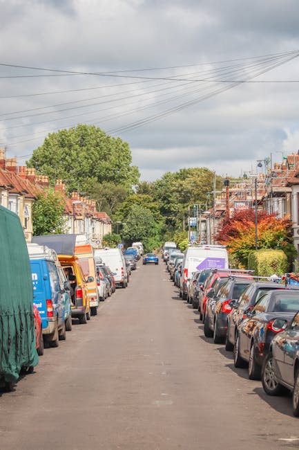 A residential street in Church End Barnet with parked cars lining both sides of the narrow tarmac road, including small hatchbacks and larger vans, some with company branding. The street features typical terraced houses with brick facades, front gardens, and some with small trees or shrubs. Overhead, multiple power lines cross the cloudy sky, which provides diffuse lighting across the scene. In the background, a large green tree and additional houses are visible at the end of the street. The image captures a calm, everyday suburban environment during daytime, relevant to house removals and local moving services provided by Finchley Man and Van, who may handle the transport and packing process involved in home relocation.