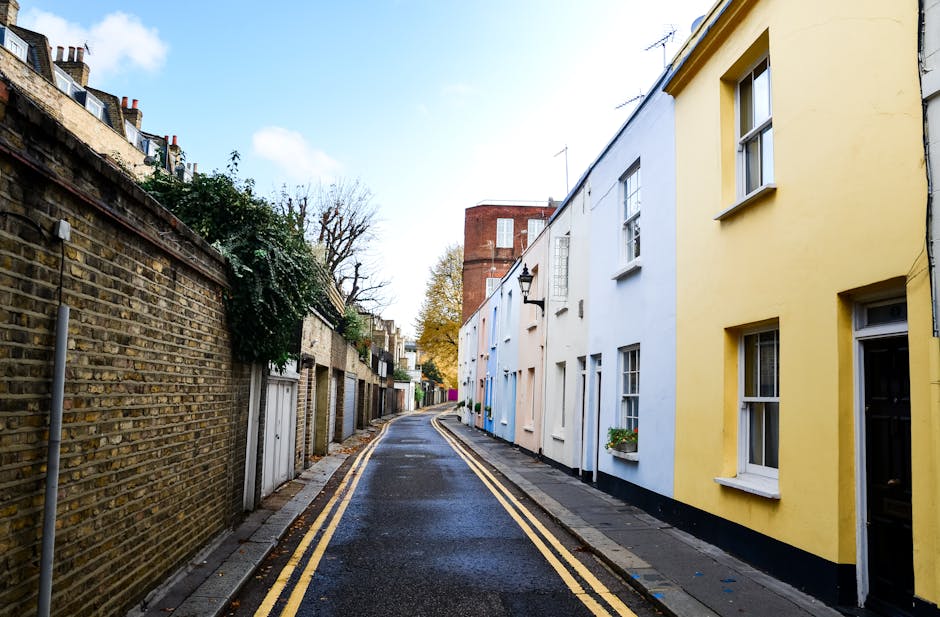 A narrow residential street with a slight curve, featuring a row of colorful terraced houses on the right side painted in pastel shades of yellow, white, and blue, each with sash windows and small flower boxes beneath. The sidewalk runs along the front of these houses, with a few outdoor lights attached to the walls. On the left side, there is an old brick wall with some greenery growing over it, separating the street from private gardens or yards. The asphalt road appears wet, possibly after rain, with double yellow lines painted along the edge indicating parking restrictions. In the distance, a taller red-brick building and a few leafless trees are visible, suggesting late autumn or winter season. The sky is partly cloudy with patches of blue, and the lighting is natural, providing a clear view of the street scene, suitable for illustrating home relocation, furniture transport, or house removals in a neighbourhood setting.