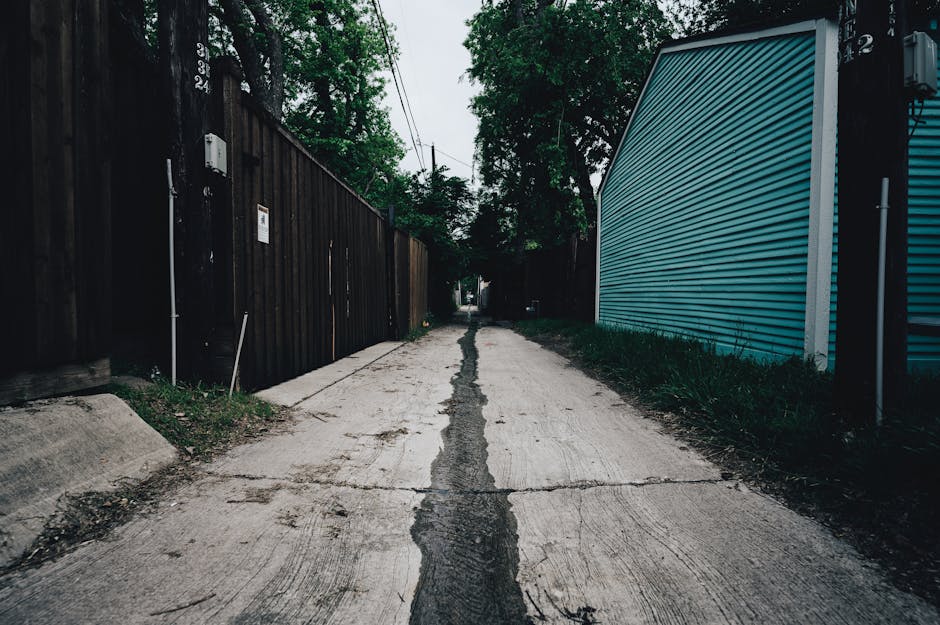 A narrow residential street with a slightly cracked concrete surface, flanked by tall wooden fencing on the left and a modern blue metal building on the right. Overhead power lines are visible above, and the street is lined with trees and greenery, creating a shaded environment. The scene appears to be a typical urban setting for house removals or furniture transport, with no vehicles or people present at the moment. The environment suggests a quiet, private area suitable for home relocation services by Finchley Man and Van, focusing on delivery or loading processes through narrow streets, with careful consideration of space and environment for effective packing and moving operations.