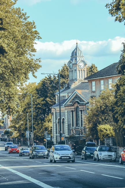 A street scene in Finchley with several parked and moving cars along the roadside, including a white van parked on the pavement. In the background, there is a historic church with a tall clock tower and a dome, surrounded by lush green trees. The sidewalk adjacent to the church has some cardboard moving boxes and packing materials, indicating recent or ongoing house removals. The area appears to be well-lit under a clear blue sky with minimal cloud cover. This setting exemplifies a typical residential and community environment where Finchley Man and Van provides home relocation and furniture transport services, assisting with packing, loading, and moving logistics within the locality.